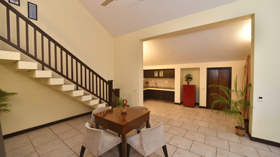 A spacious villa living area at Vedic Village Spa Resort featuring a wooden staircase tiled flooring a dining table ceiling fan and warm ambient lighting in an open layout.