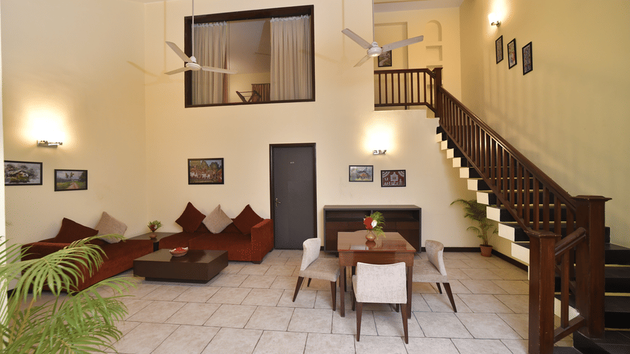 A villa lounge area at Vedic Village Spa Resort featuring low cushioned seating a wooden coffee table tiled flooring staircase and warm ambient lighting with indoor plants.
