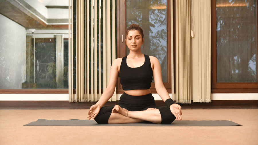 A woman in black workout attire sitting in a meditation pose on a yoga mat in a spacious studio at Vedic Village Spa Resort with large windows and warm ambient ceiling lighting.