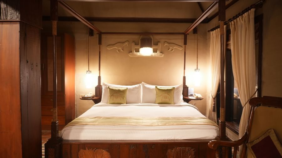 Elegantly styled four-poster king size bed in the Zamindar Suite at Vedic Village Spa Resort, complemented by a wooden lounge chair, and soft lighting.