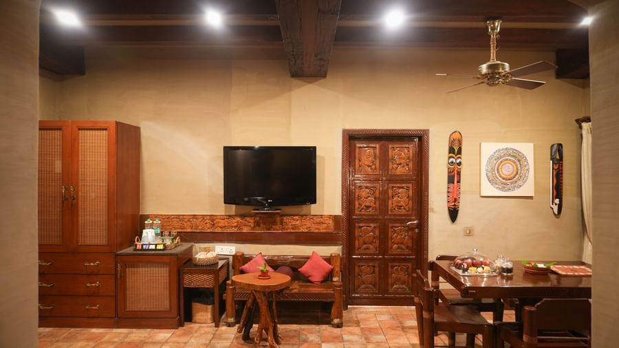 Living area of the Zamindar Suite at Vedic Village Spa Resort with an in-room dining setup, warm ambient lighting, wooden furnishings, and a wall-mounted TV.