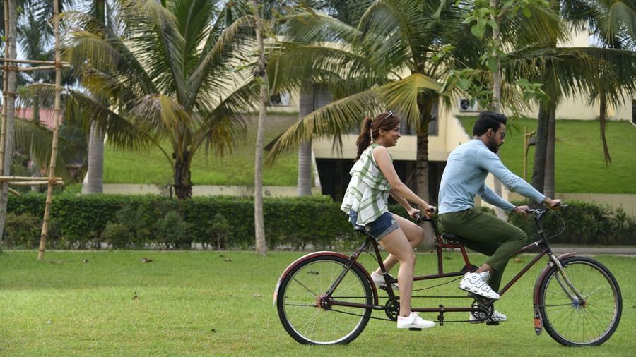 A couple rides a tandem bicycle on a grassy lawn with palm trees in the background
