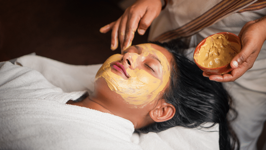 Female guest lying on a treatment table with a golden turmeric face mask applied during an ayurvedic facial therapy session at Vedic Village Spa Resort Kolkata.