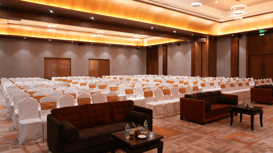 Empty banquet hall at Vedic Village Spa Resort with wooden wall panelling, ornate ceiling lighting, a dark sofa seating area, and a grand piano in the corner.
