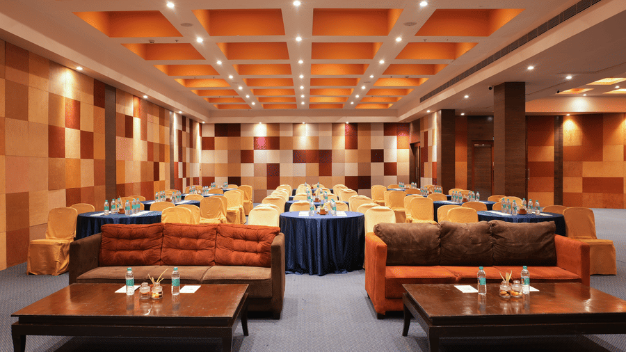  Banquet lounge hall at Vedic Village Spa Resort Kolkata with low seating sofas, round tables with blue linen, black chairs, and a warm coffered ceiling with decorative lighting.
