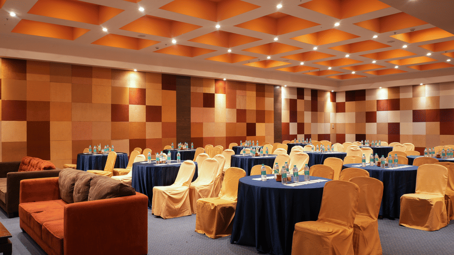 Banquet hall at Vedic Village Spa Resort Kolkata featuring round tables with blue linen, orange sofa seating, black chairs, and a warm coffered ceiling with recessed lighting.