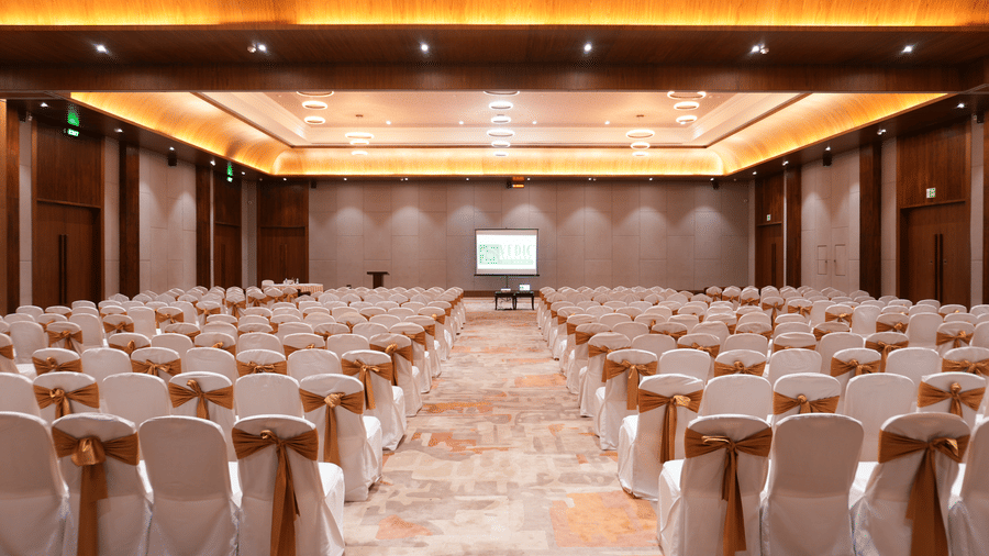 Large banquet hall at Vedic Village Spa Resort arranged in theatre style with rows of white chair-covered seating, a projection screen, and warm wooden ceiling lighting.