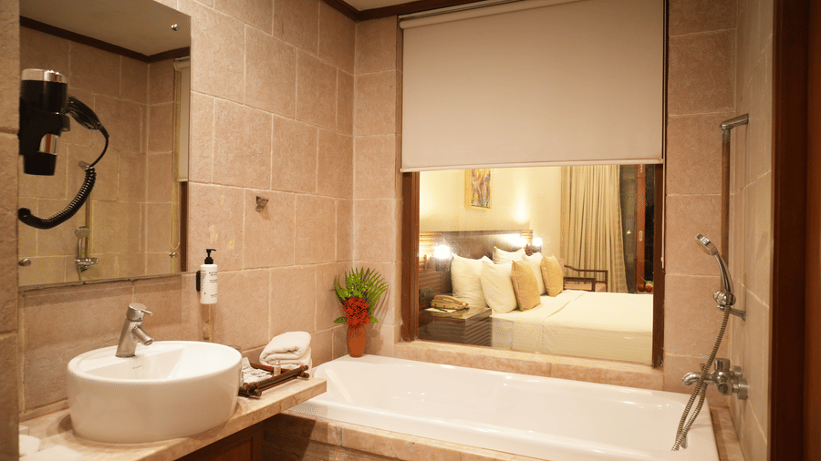 Washroom of the Standard Suite at Vedic Village Spa Resort featuring a sink, wall mirror, and a bathtub set against a glass-panelled wall overlooking the room, with towels and amenities placed beside the sink.