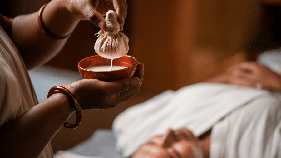 Therapist applying a herbal pouch kizhi treatment on the face of a female guest lying on a treatment table at Vedic Village Spa Resort Kolkata.