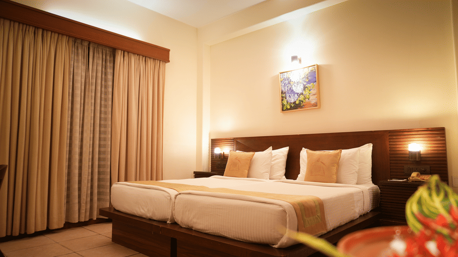 A warmly lit Superior Room at Vedic Village Spa Resort featuring a double bed with a wooden headrest, drapes over a large window, and a painting above the bed.