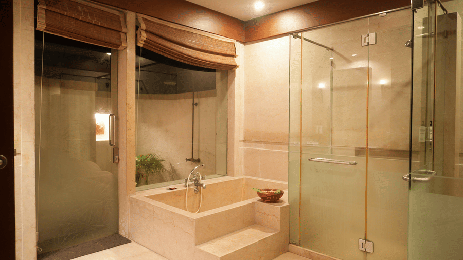 Glass-panelled shower room in the Lake View Villa at Vedic Village Spa Resort with a bathtub, shower cubicle, and a glass door opening to an outdoor shower area.