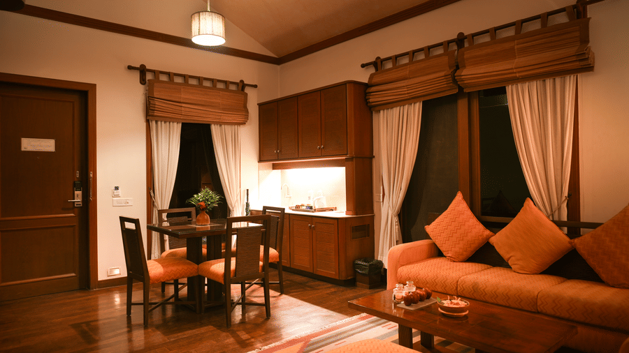 Warmly lit living area in the Lake View Villa at Vedic Village Spa Resort with in-room dining space, carpeted floors, plush couch, and wooden furnishings.