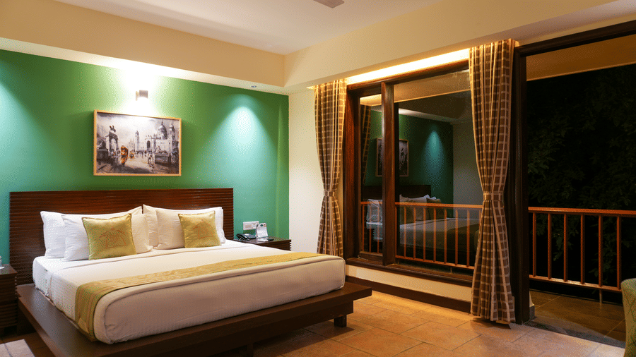 Villa bedroom at Vedic Village Spa Resort Kolkata featuring a king bed, bold green accent wall, framed artwork, ceiling fan, and large balcony doors with natural light.