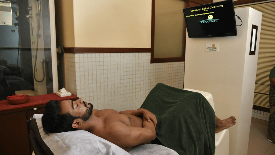 Male guest lying on a treatment table receiving an ayurvedic body therapy session in a warmly lit treatment room at Vedic Village Spa Resort.