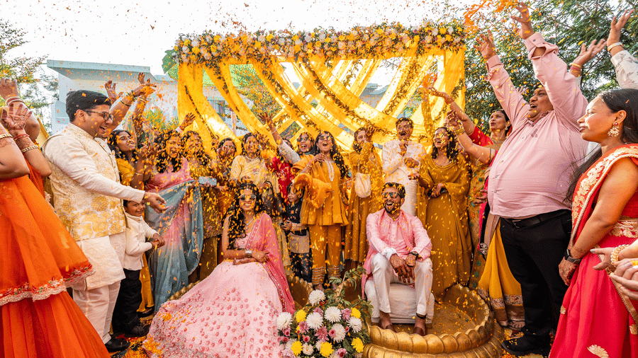 A vibrant moment from a Haldi ceremony where family members shower the couple sitting under a yellow and orange floral canopy with flower petals at Vedikant Resorts The Mallard Corbett.