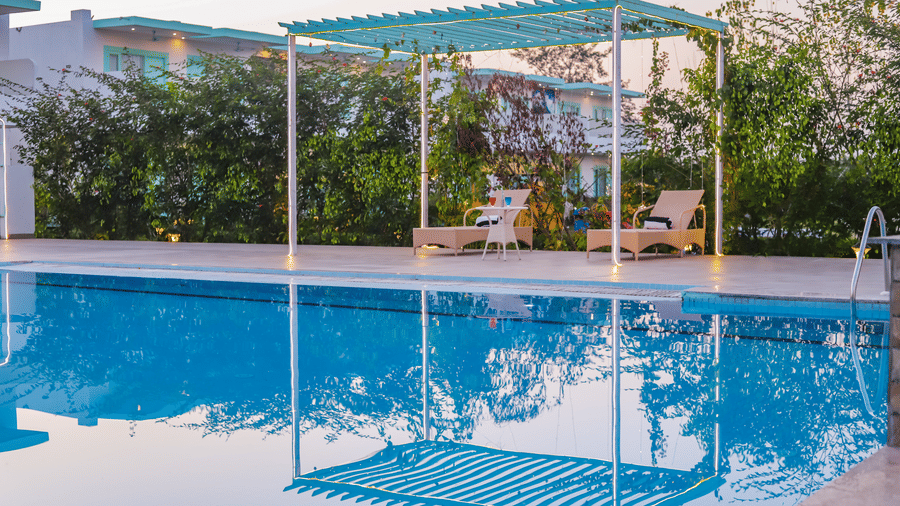 View of a swimming pool with buildings, trees and clear blue sky in the background - Vedikant Resorts The Mallard Corbett.