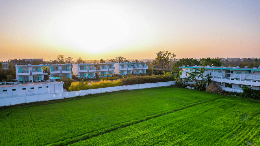 Aerial view of Vedikant Resorts The Mallard Corbett with the building complex, open fields, and trees under a setting sky.