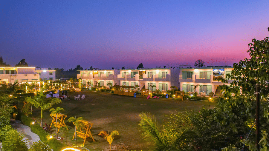 Wide-angle evening view of Vedikant Resorts The Mallard Corbett showing rooms and outdoor lawn with scattered tables and chairs.