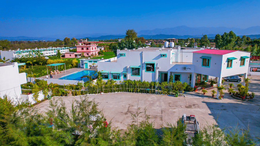 An exterior view of Vedikant Resorts The Mallard Corbett shows buildings, a pool, and surrounding landscape under a blue sky.