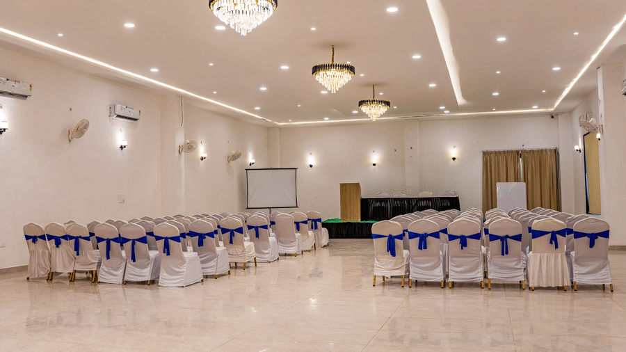 Banquet hall at Vedikant Resorts, The Mallard Corbett featuring seating area with chairs facing towards the centre of the stage which has a table and chairs. The entire space is equipped with chandeliers, ceiling lights, AC, whiteboard, white screen, and m