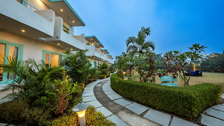 Exterior view of rooms at Vedikant Resorts The Mallard Corbett featuring a pavement filled with trees on either side and a lawn, and two storey buildings consisting of diverse accommodation. 
