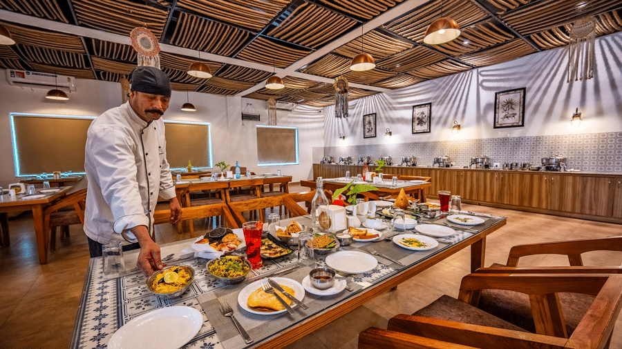 The restaurant staff is placing food at the table in Himalayan Spices at Vedikant Resorts The Mallard Corbett. The table also showcases several other dishes surrounded by chairs, and there are other sets of dining tables and chairs as well.