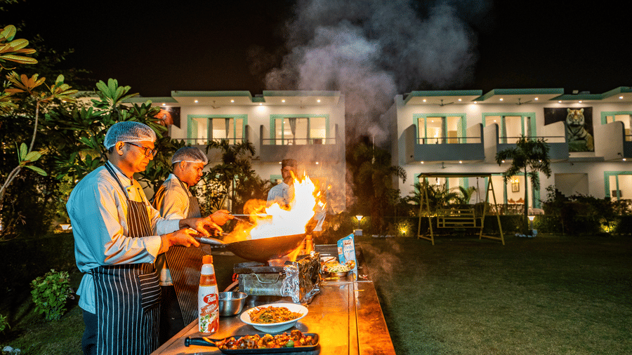 Outdoor cooking at Vedikant Resorts The Mallard Corbett featuring three chefs cooking near a lawn where there is a set up of tables and chairs near the buildings of the property.