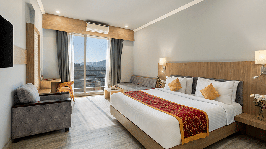 A hotel room with a bed, seating area, desk, and view through a large window - Via Lakhela Resort & Spa, Kumbhalgarh