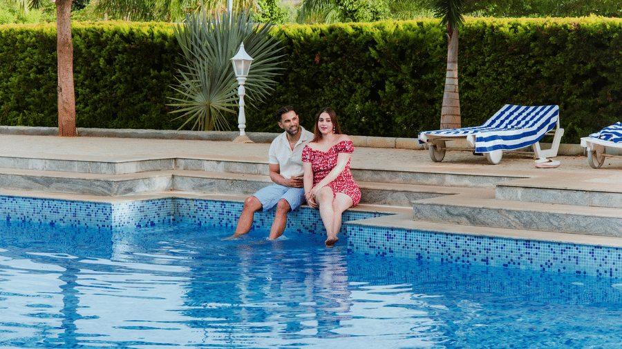 A man and a woman sitting on the edge of a blue outdoor swimming pool with their feet in the water | Vintage Trails By Ilara