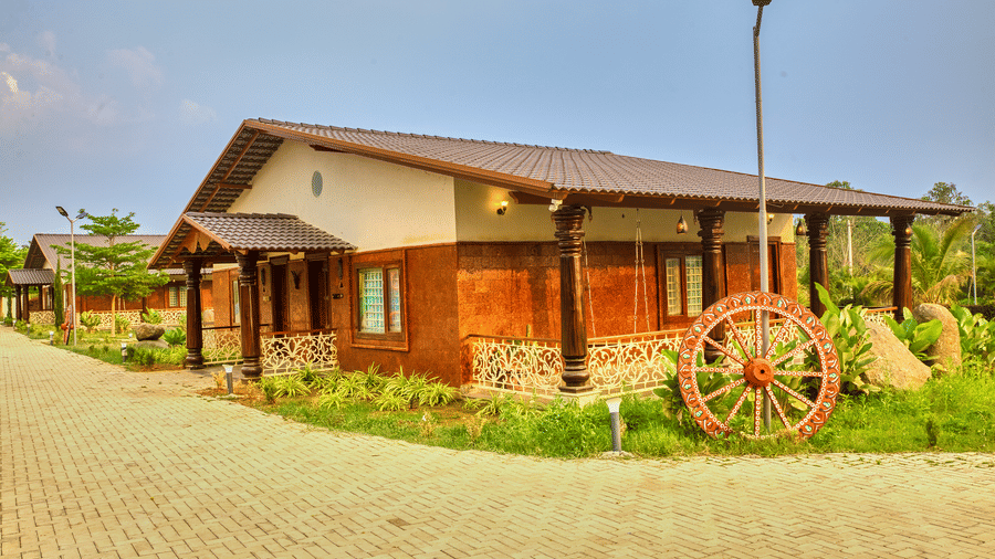 The exterior of a single-storey building with a sloping roof and wooden accents, situated on a paved path with a decorative wagon wheel nearby. - Vintage Trails by Ilara