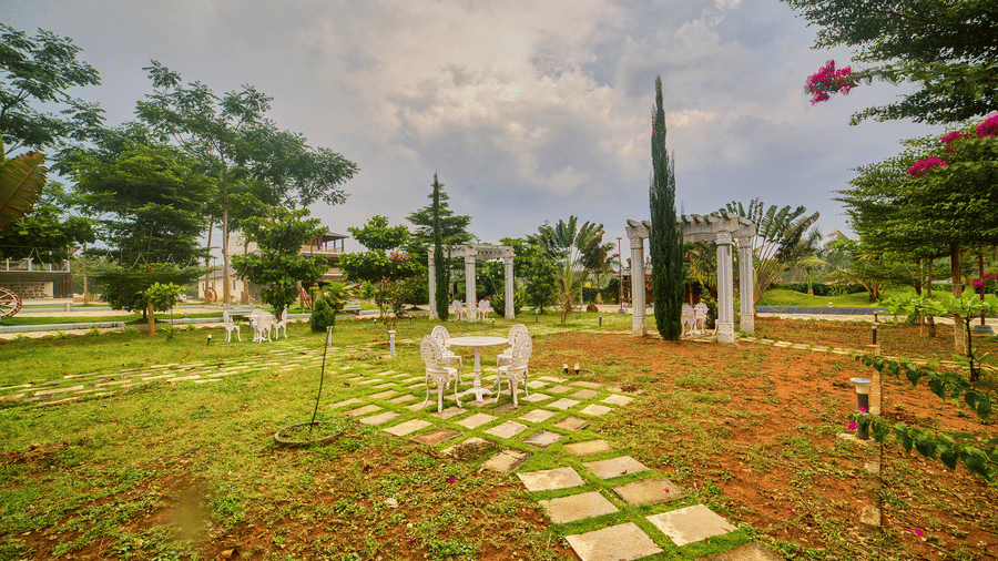 An expansive garden featuring white wrought-iron tables and chairs on stone paths, classical pergolas, and manicured trees under a dramatic, cloudy sky - Vintage Trails By Ilara, Sakleshpur