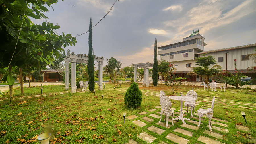 A wide outdoor view of a green lawn with a small table and chairs set, leading towards a modern building under a cloudy sky - Vintage Trails By Ilara