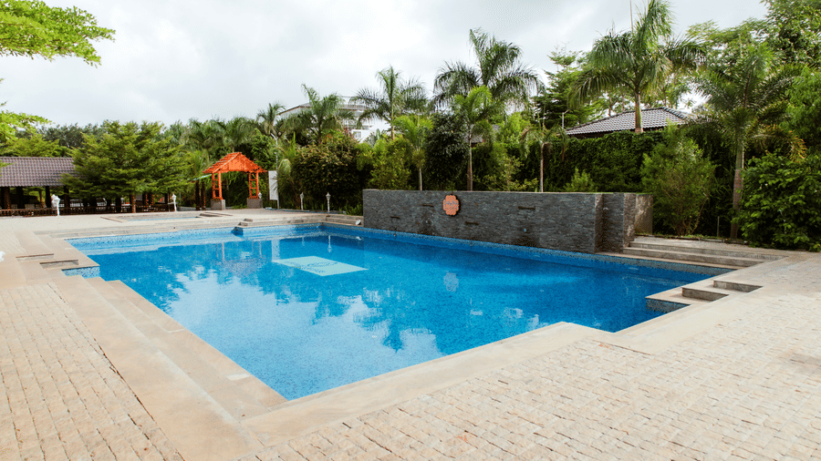 A wide view of an outdoor blue swimming pool surrounded by paved tiling, trees, and hedges | Vintage Trails By Ilara