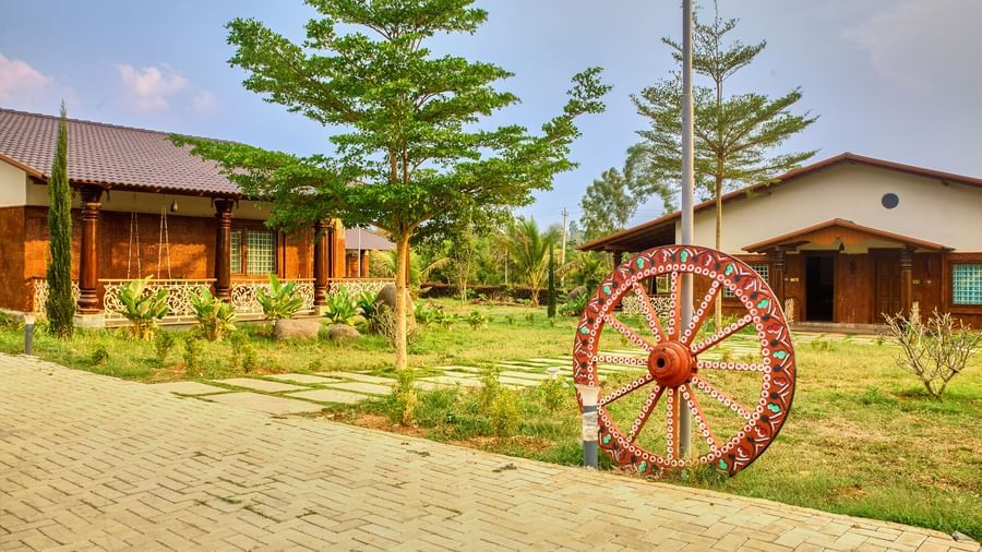 The exterior of a single-storey building with a sloping roof and wooden accents, situated on a paved path with a large decorative red wagon wheel in the foreground. - Vintage Trails by Ilara