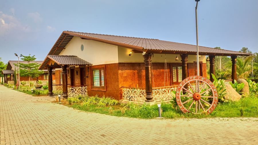 The exterior of a single-storey building with a sloping roof and wooden accents, situated on a paved path with a decorative wagon wheel nearby. - Vintage Trails by Ilara