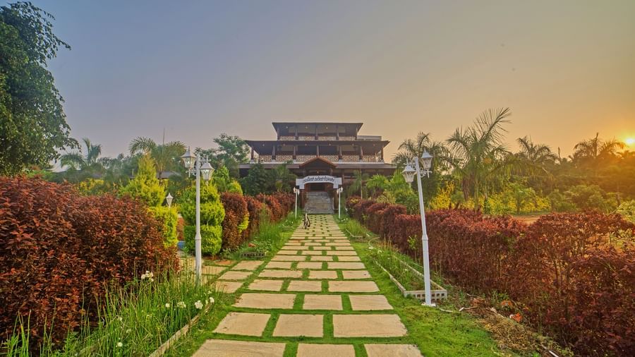 A long pathway of square stepping stones through a vibrant green garden, leading towards a grand, traditional building with multi-tiered roofs under a sunset sky. - Vintage Trails by Ilara