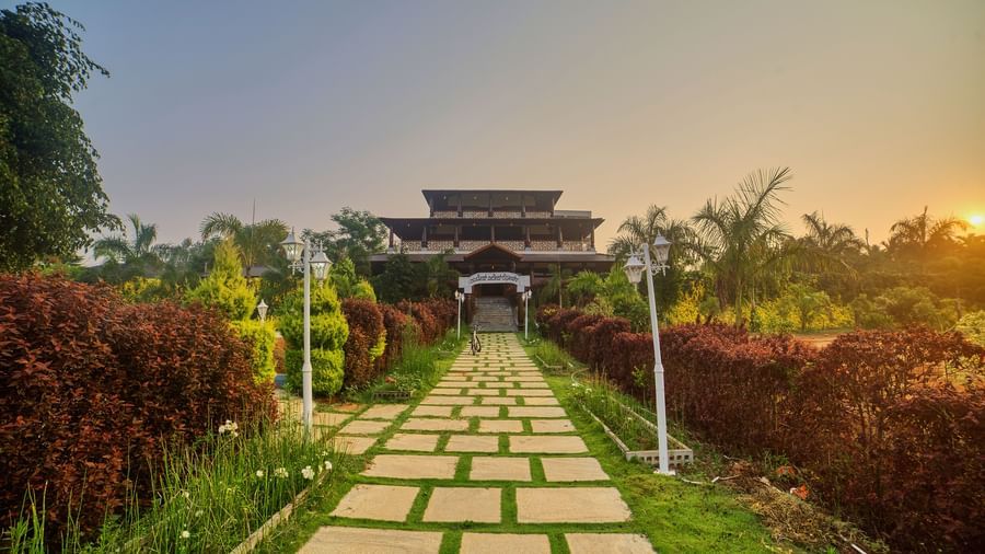  A pathway made of square stepping stones leading through a well-maintained green garden towards a grand, possibly ancient or traditional, entrance structure with lush trees on either side. - Vintage Trails by Ilara