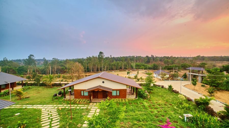 A ground-level view of a single cottage with a dark roof and warm-toned walls, surrounded by a green lawn and a paved pathway, under a vivid sunset sky - Vintage Trails by Ilara