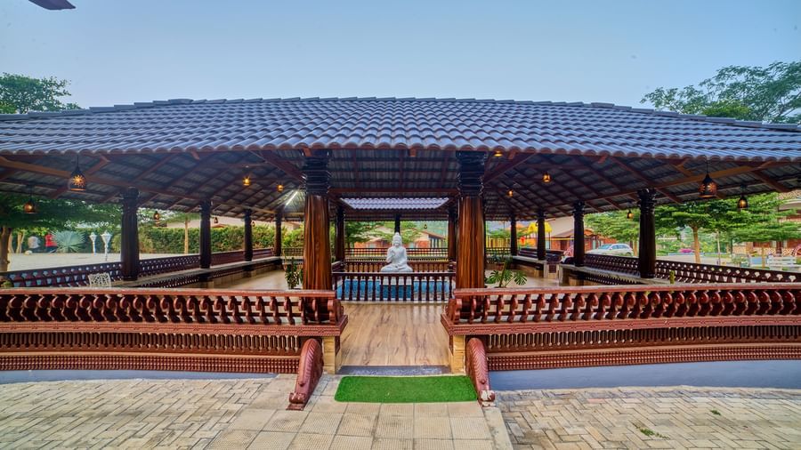 An open-sided wooden pavilion with a dark tiled roof and built-in seating, situated on a paved area, under a clear blue sky. - Vintage Trails by Ilara