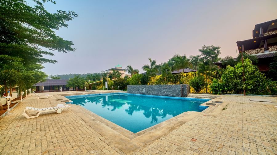 An outdoor swimming pool with blue water and white sun loungers on the paved surround, with a traditional-style building and lush trees in the background under a sunset sky, from a slightly different angle. - Vintage Trails by Ilara