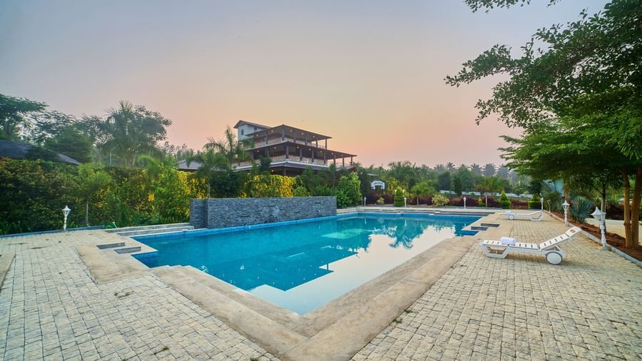 An outdoor swimming pool with blue water and white sun loungers on the paved surround, with a traditional-style building and lush trees in the background under a sunset sky. - Vintage Trails by Ilara