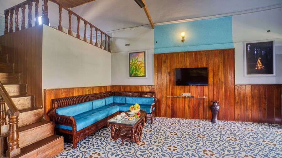 The interior of a spacious living area with patterned tiled floor, featuring a blue curved sofa, a wooden TV stand, and stairs leading to an upper level.