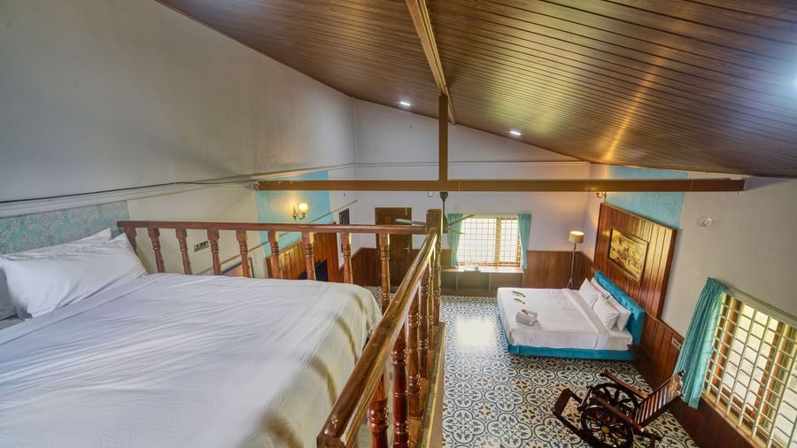 A loft bedroom with a large white bed on an elevated wooden platform, a pitched wooden ceiling, and a sitting area with a rocking chair visible below, all within a brightly lit room.