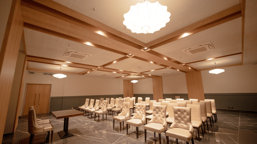 The well-lit event space at Vinti Resort, Ambaji, features wooden beams on the ceiling and neat rows of comfortable, high-backed chairs.