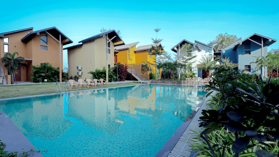 The sparkling swimming pool at Vinti Resort, Ambaji, is framed by brightly coloured, modern individual resort cottages and lush foliage.