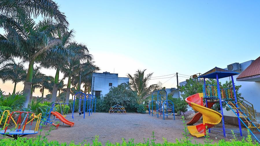 A vibrant outdoor children's play area with swings and slides, surrounded by green grass and palm trees - Vishal Lords Inn Gir Forest