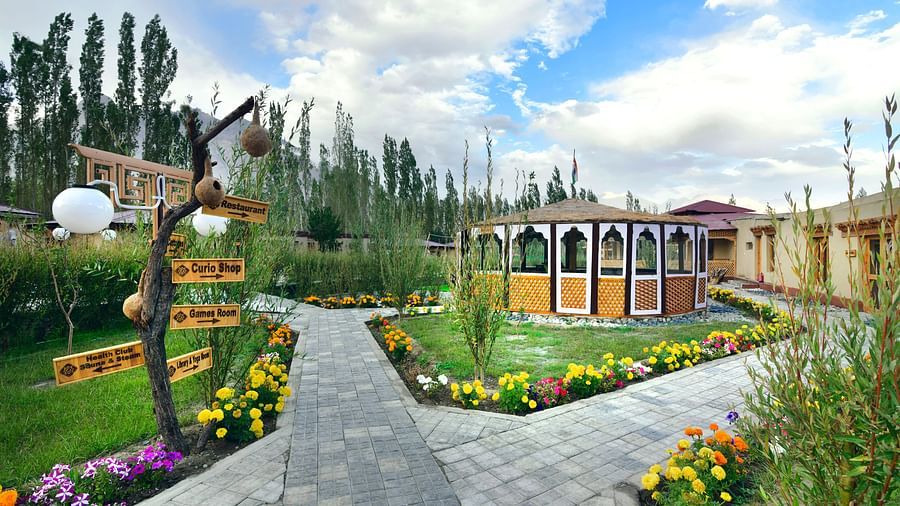 A pathway leading to White Sands Resort, Ladakh lined with flowers and greenery, with decorative signage and accommodation buildings visible against a backdrop of trees and a cloudy sky.