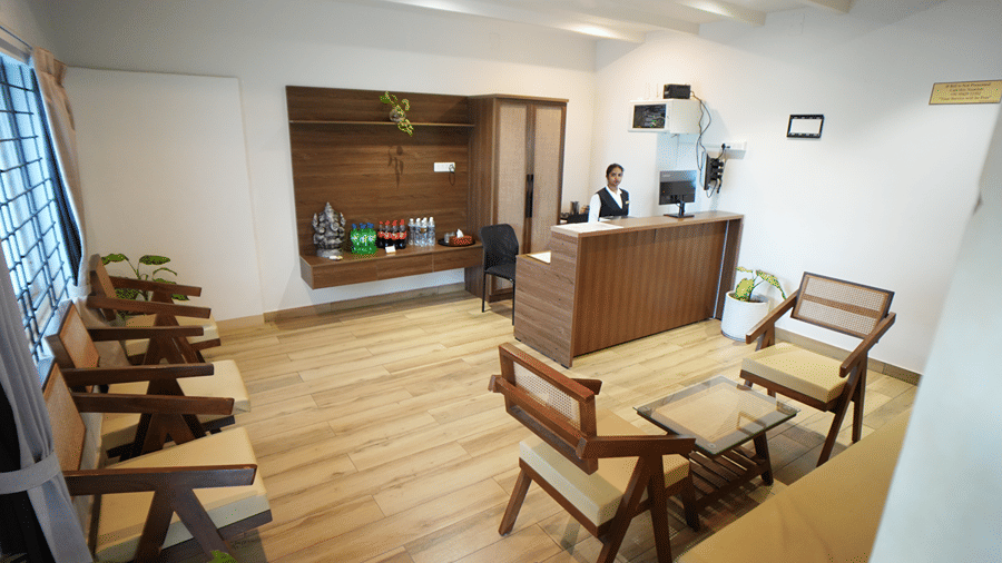 An indoor reception area featuring a front desk with a staff member standing behind it, wooden flooring, and a small seating arrangement with chairs and a table.
