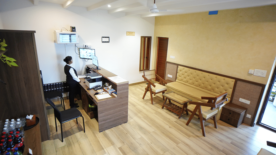 An indoor reception area featuring a front desk with a staff member standing behind it, wooden flooring, and a small seating arrangement with chairs and a table.