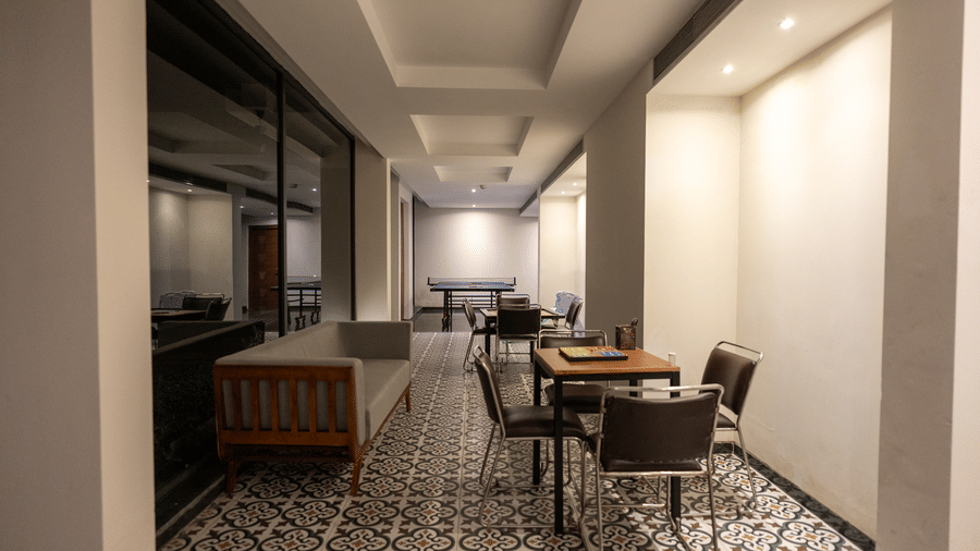 A modern lobby-style acitivity room at Wyndham Garden featuring a patterned tile floor, a long wooden sofa on the left, small dining tables with chairs on the right, ceiling lighting, plain walls and columns, and a large reflective glass wall.